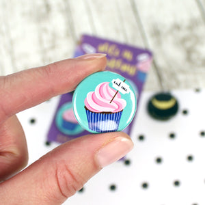 A cupcake in a blue paper case with pink frosting and an ‘eat me’ label